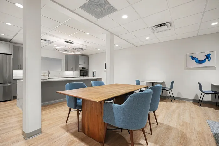 Modern dining area with elegant wooden table, surrounded by blue chairs, leads to a sleek kitchen with stainless steel appliances and contemporary cabinetry.