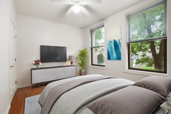 Inviting bedroom with a stylish decor, featuring large windows offering natural light, a cozy bed with plush bedding, a modern TV setup, and elegant artwork complementing the serene ambiance.