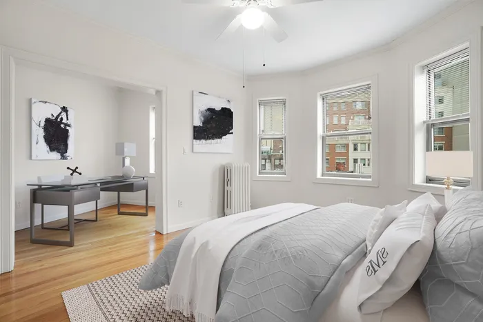 Bright and inviting bedroom featuring elegant white bedding, three large windows with city views, and a chic adjoining office space.