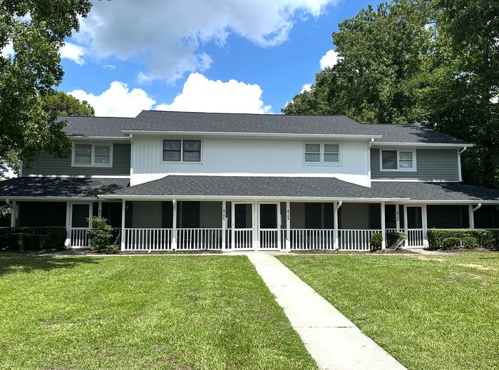 Charming two-story townhouse with a spacious front porch, surrounded by lush greenery and tall trees under a clear blue sky.
