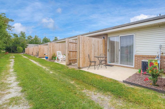 Charming outdoor patio space with wooden privacy fences, cozy seating area, and lush greenery under a bright blue sky.