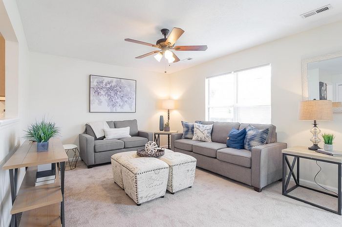 Inviting living room with cozy gray sofas, stylish ottomans, modern ceiling fan, and elegant decor, offering a warm and welcoming atmosphere.