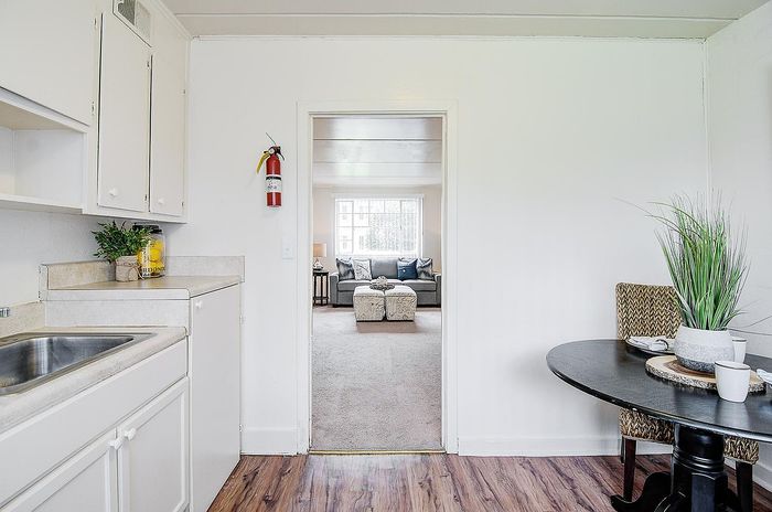 Cozy apartment kitchen with modern white cabinets, leading to a bright living room featuring a stylish gray sofa and accent chairs, perfect for relaxation.