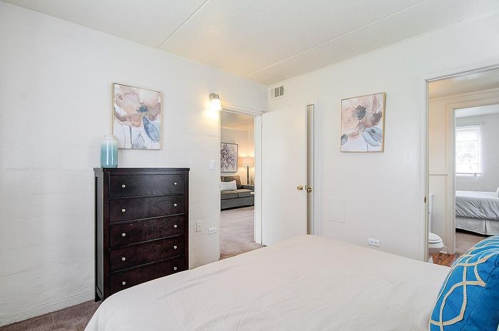Bright bedroom with modern decor, featuring a dark wood dresser, elegant wall art, and a glimpse into a cozy living space.