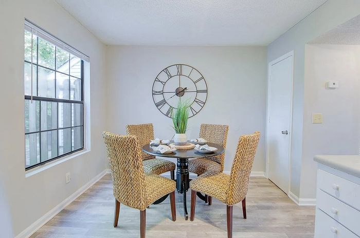 Charming dining area with woven chairs, elegant wall clock, and large window view in a bright and welcoming apartment setting.