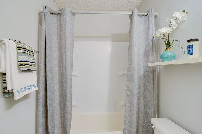Bright and modern bathroom with a clean shower area, elegant grey curtains, fresh white towels, and decorative touches like a vase with flowers on a floating shelf.