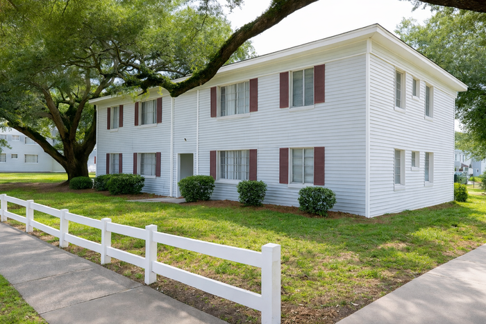 Charming two-story white apartment building with red shutters, surrounded by lush greenery and a white picket fence.