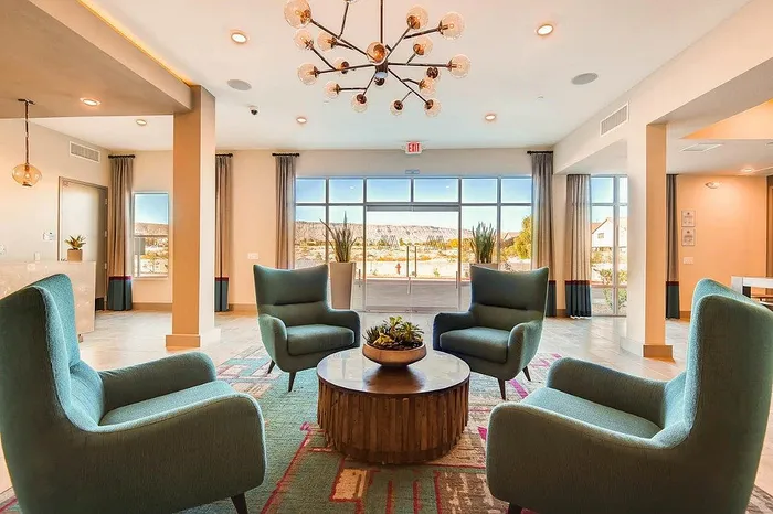 Elegant apartment lounge with plush teal armchairs, a modern chandelier, and abundant natural light from large windows.