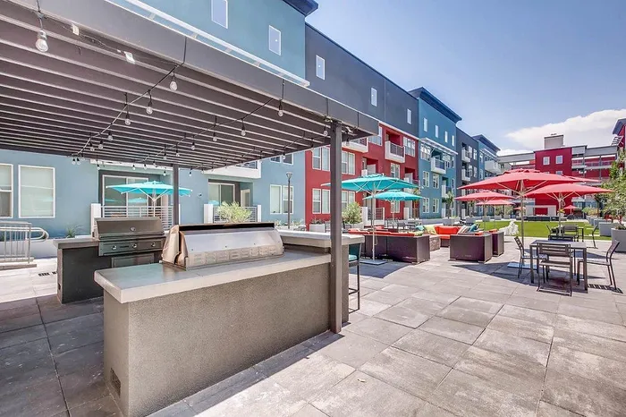 Modern apartment community courtyard with sleek grill station, vibrant red and turquoise umbrellas, and stylish outdoor seating for social gatherings.