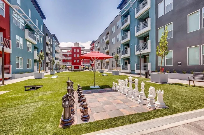 Vibrant apartment courtyard with colorful buildings, lush green lawn, outdoor games, and giant chess set under a sunny sky.