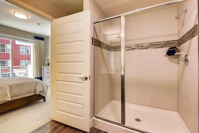 Stylishly modern apartment bathroom with a sleek glass-enclosed shower, elegantly transitioning to a cozy bedroom with ample natural lighting.