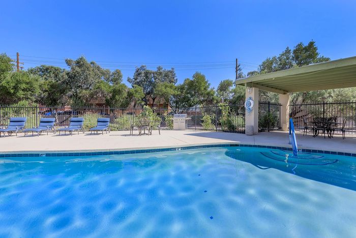 Inviting outdoor pool area with lounge chairs and a shaded patio, surrounded by lush greenery and clear blue skies.