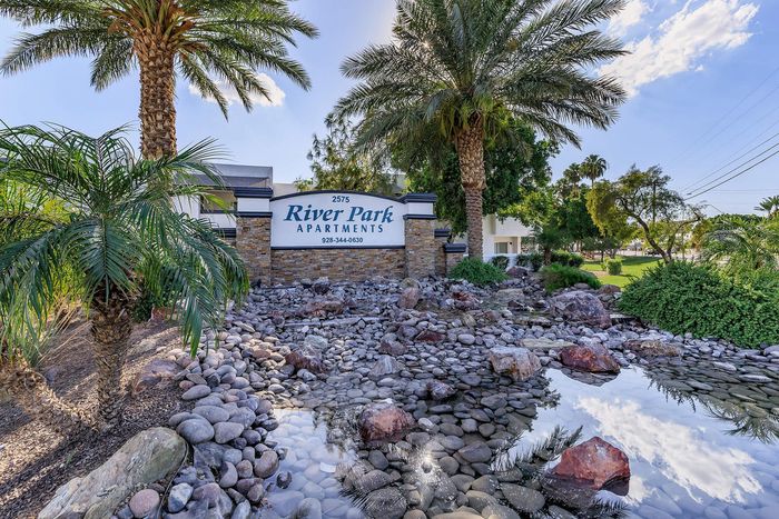 Scenic entrance of River Park Apartments featuring lush palm trees, a serene rock garden, and tranquil water reflection.