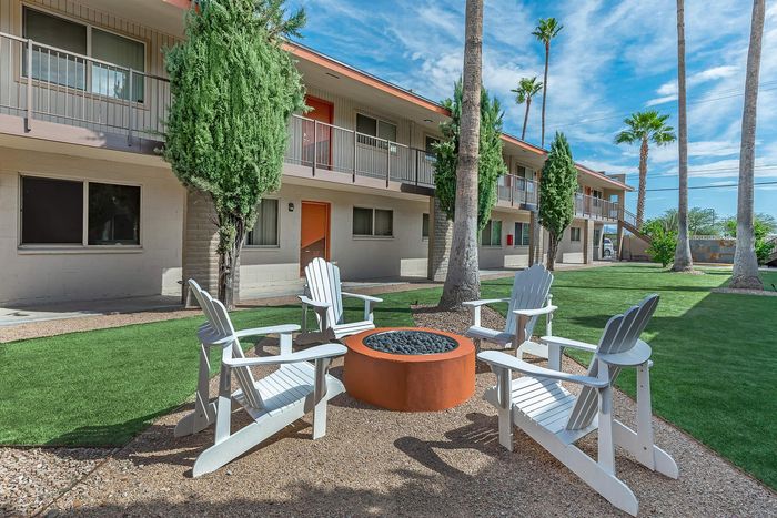 Charming outdoor courtyard with cozy fire pit surrounded by white Adirondack chairs, lush greenery, and modern apartment buildings under a clear blue sky.