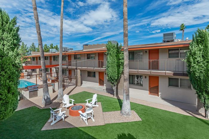 Charming apartment exterior showcasing a lush green courtyard with inviting Adirondack chairs around a fire pit, and a shimmering pool in the background under a bright blue sky.