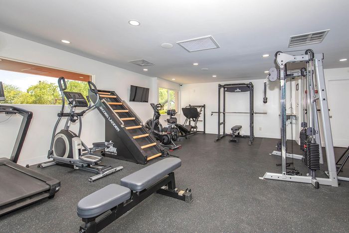 Modern fitness center in luxury apartment with state-of-the-art exercise equipment, including treadmill, elliptical, and weight machines, set in a bright and spacious room with large windows.