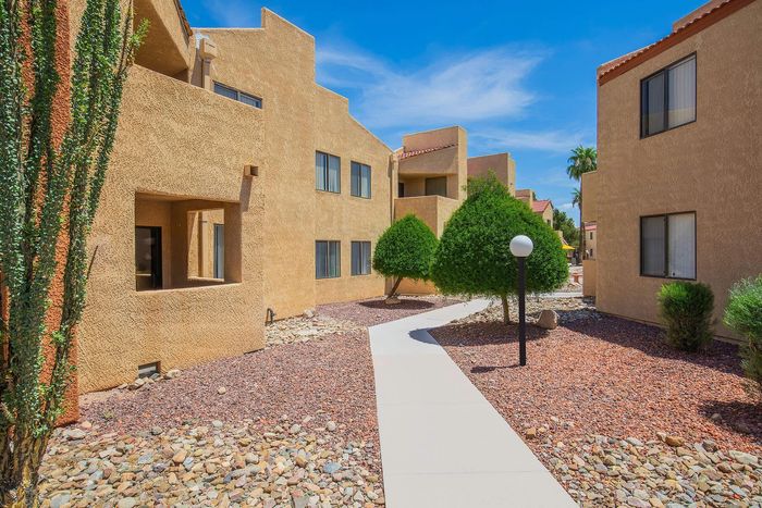 Desert oasis-style apartment complex with charming stucco exteriors, vibrant desert landscaping, and a serene walkway under blue skies.