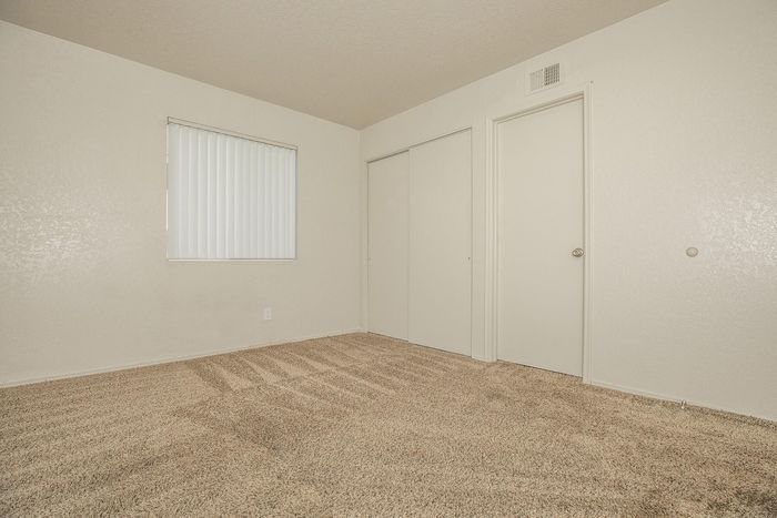 Bright and inviting bedroom with soft beige carpeting, white walls, and ample natural light through a window with vertical blinds. Features a convenient closet and door, offering a versatile and comfortable living space.