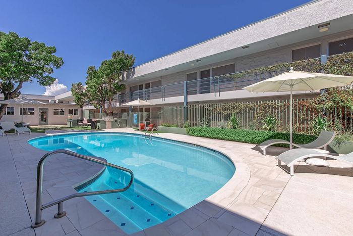 Inviting apartment complex courtyard featuring a sparkling swimming pool, lounge chairs, and vibrant greenery under a clear blue sky.