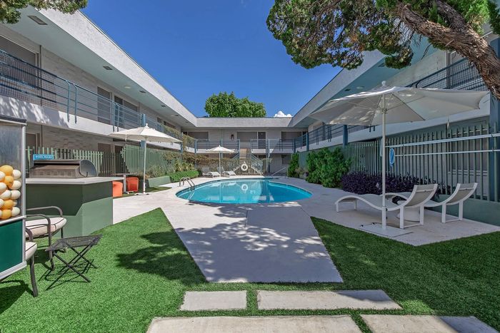 Inviting apartment courtyard with a sparkling pool, sun loungers, and modern outdoor seating under clear blue skies.
