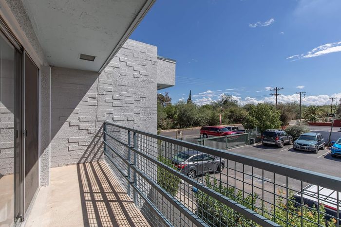 Sunlit balcony with wrought iron railing offering serene views of a landscaped parking area and lush greenery.