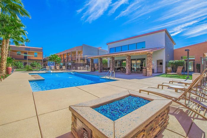 Inviting apartment complex featuring a sparkling pool with lounging chairs, palm trees, and modern architecture under a clear blue sky.