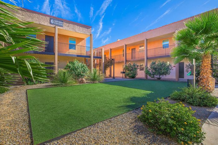 Charming apartment complex featuring vibrant landscaping with lush greenery and a spacious courtyard, set against a clear blue sky.