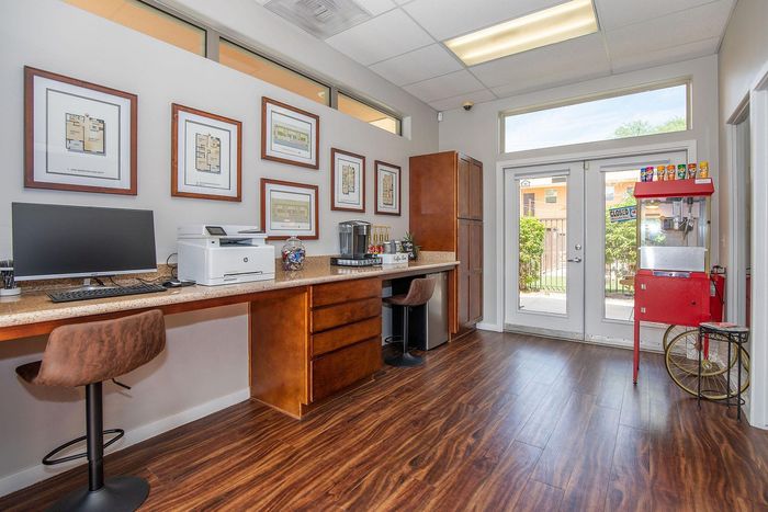 Chic and functional workspace with wood flooring, featuring a modern desk setup, wall-mounted art, and a convenient refreshment station.