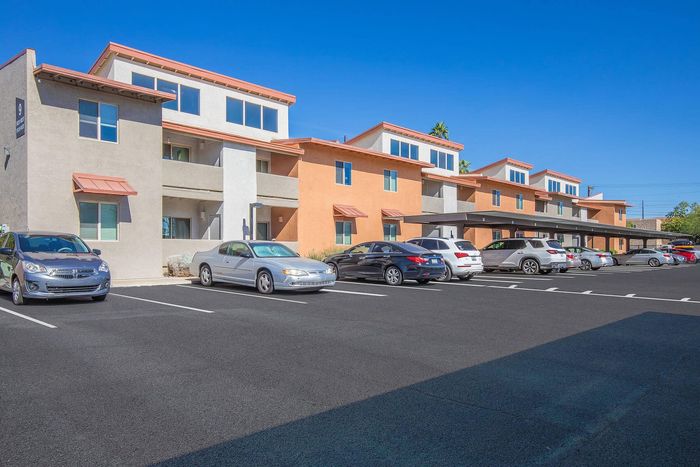 Modern apartment complex with vibrant orange accents, ample parking, and sunlit windows under a clear blue sky.