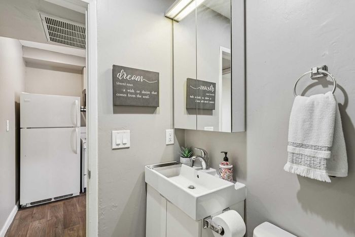 Charming apartment bathroom featuring a sleek white vanity with mirror, modern lighting, and a subtle decorative touch with a potted plant. Conveniently adjacent to a bright kitchen space with a large refrigerator.