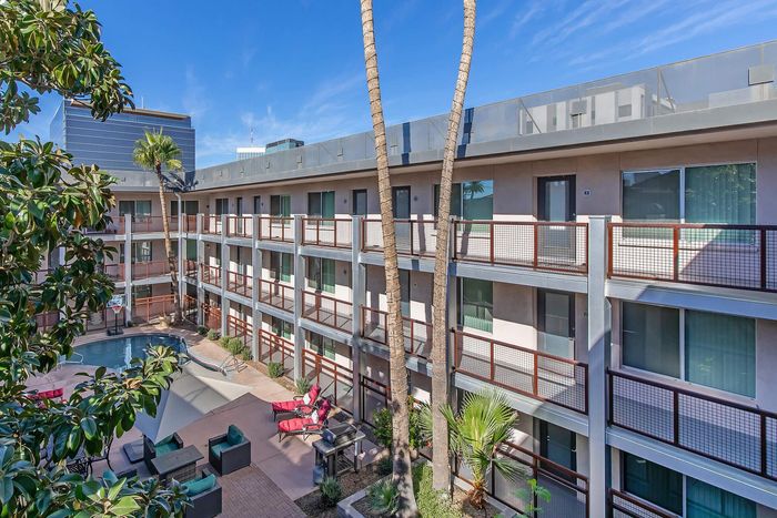 Modern apartment complex featuring a lush courtyard with a swimming pool, vibrant outdoor lounge furniture, and palm trees under a clear blue sky.