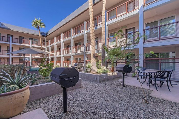 Modern apartment courtyard featuring a stylish patio area with BBQ grills, lush landscaping, and elegant seating under a vibrant blue sky.