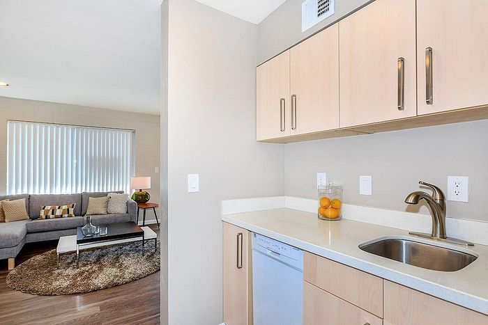 Modern apartment corner showcasing a cozy living room with a sleek gray sofa, a stylish side table, and a well-appointed kitchen space with elegant cabinetry and appliances.