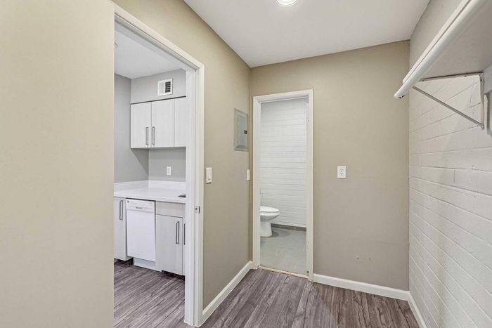 Modern apartment hallway leading to a bright kitchen with sleek white cabinetry and a well-appointed bathroom with stylish brick accents.