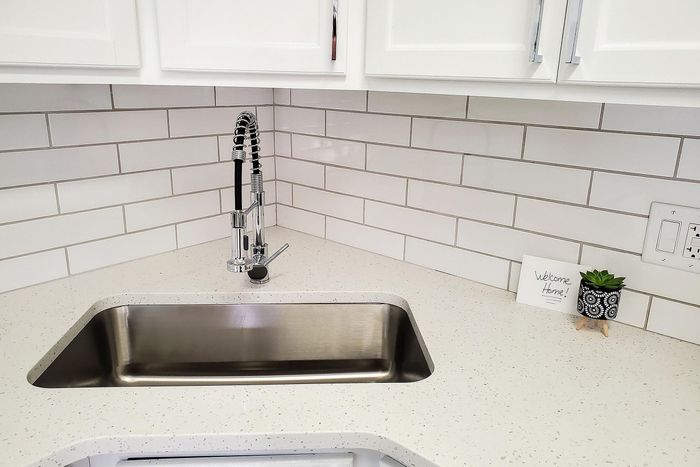 Modern kitchen corner featuring sleek stainless steel sink, elegant white tile backsplash, and a cheerful 'Welcome Home' card next to a stylish plant.