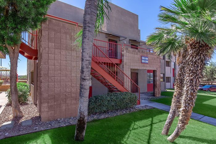 Charming apartment exterior with modern red railings, surrounded by lush greenery and palm trees under a bright blue sky.