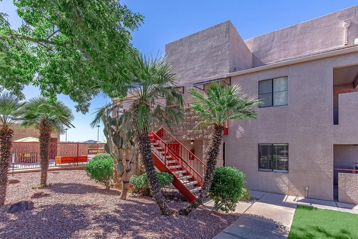 Charming apartment exterior with vibrant landscaping, palm trees, and a welcoming red staircase, all under a clear blue sky.