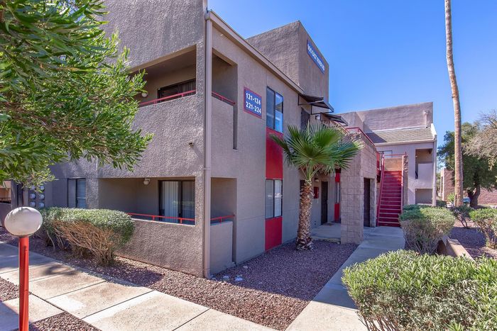 Charming apartment exterior with modern design, featuring red accents, manicured greenery, and a sunlit pathway.