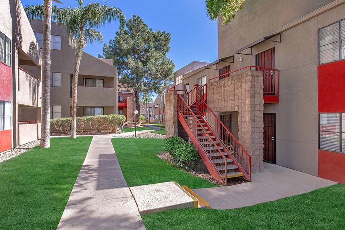Inviting apartment exterior with vibrant red accents, lush greenery, and palm trees under a clear blue sky.