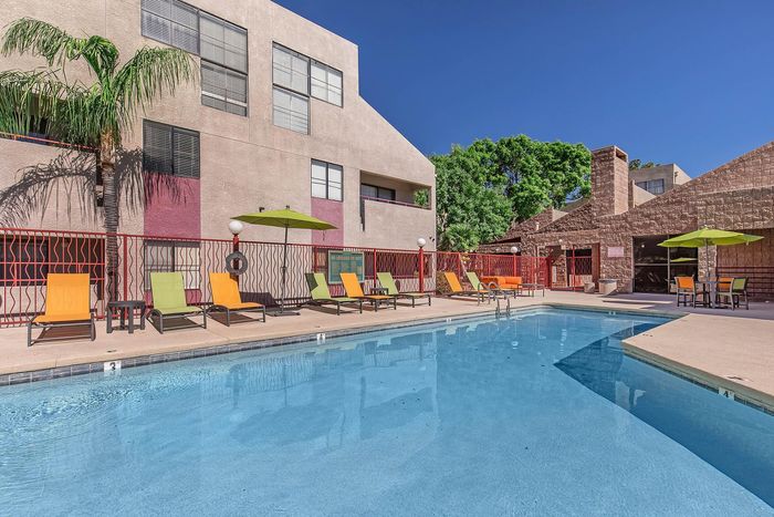 Vibrant apartment pool area with colorful lounge chairs, lush greenery, and a relaxing ambiance under bright blue skies.