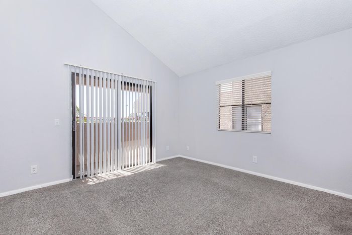 Bright and airy bedroom with cozy carpet flooring, vertical blinds leading to a balcony, and large windows offering natural light.