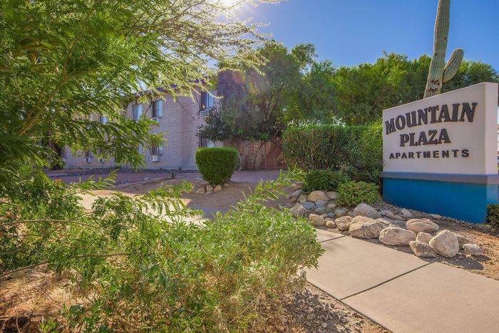 Inviting entrance to Mountain Plaza Apartments, surrounded by lush greenery and bright desert landscaping under a clear blue sky.