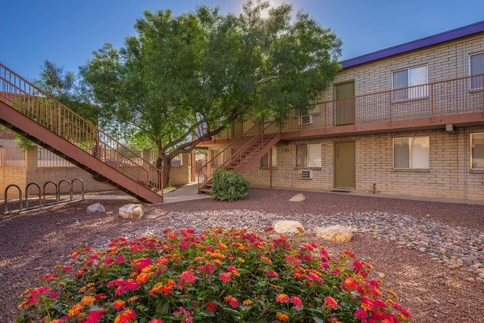 Charming two-story apartment complex with lush green trees and vibrant flower garden, featuring rustic exterior and inviting pathways.