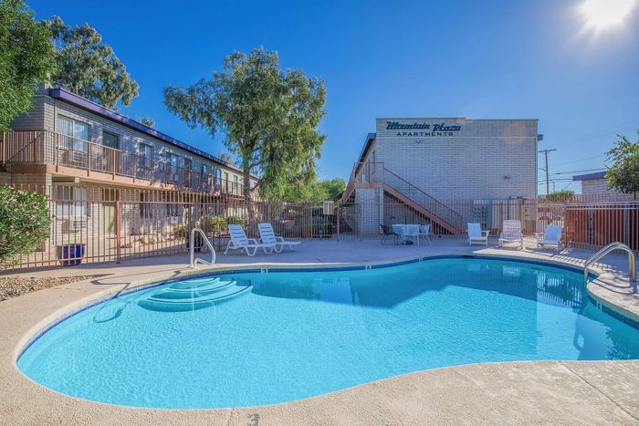 Inviting apartment complex featuring a crystal-clear swimming pool, surrounded by lush greenery and sunlit patio seating.
