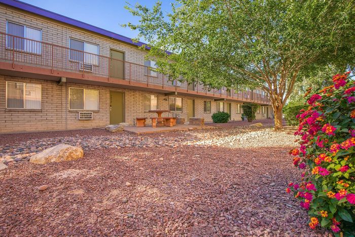 Charming apartment complex exterior with lush greenery, vibrant flowers, and inviting picnic benches beneath the shade of a large tree.