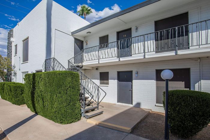Charming white brick apartment building with modern railings and lush green hedges, set under a bright blue sky.