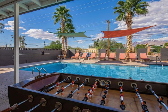 Chic outdoor pool area with lounge chairs, vibrant orange sun shades, and a foosball table under palm trees, perfect for relaxation and fun.