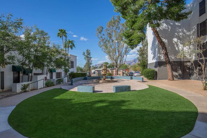 Elegant outdoor courtyard surrounded by lush greenery and modern buildings, featuring a classic stone fountain under a clear blue sky.