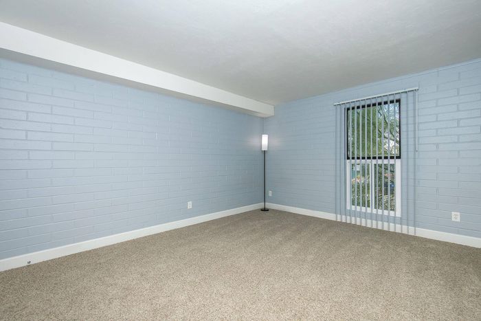 Bright and airy room with sleek modern brick walls, plush carpet, and a large window with vertical blinds, perfect for cozy living.