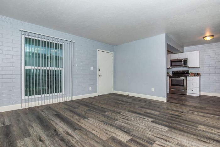 Modern apartment interior featuring sleek hardwood floors, a bright window with vertical blinds, and a stylish kitchen equipped with stainless steel appliances.